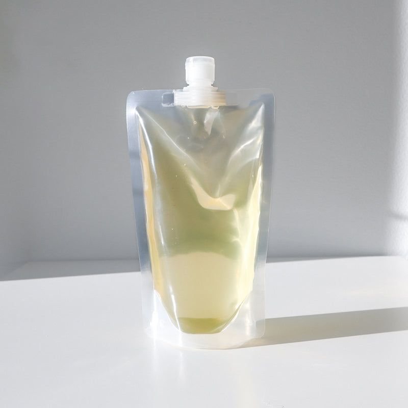 A clear stand-up pouch with a spout cap, filled with light yellow Hair, Beard & Body Oil by Green Life Trading Co., featuring nourishing jojoba oil, rests on a white surface against a light gray background.