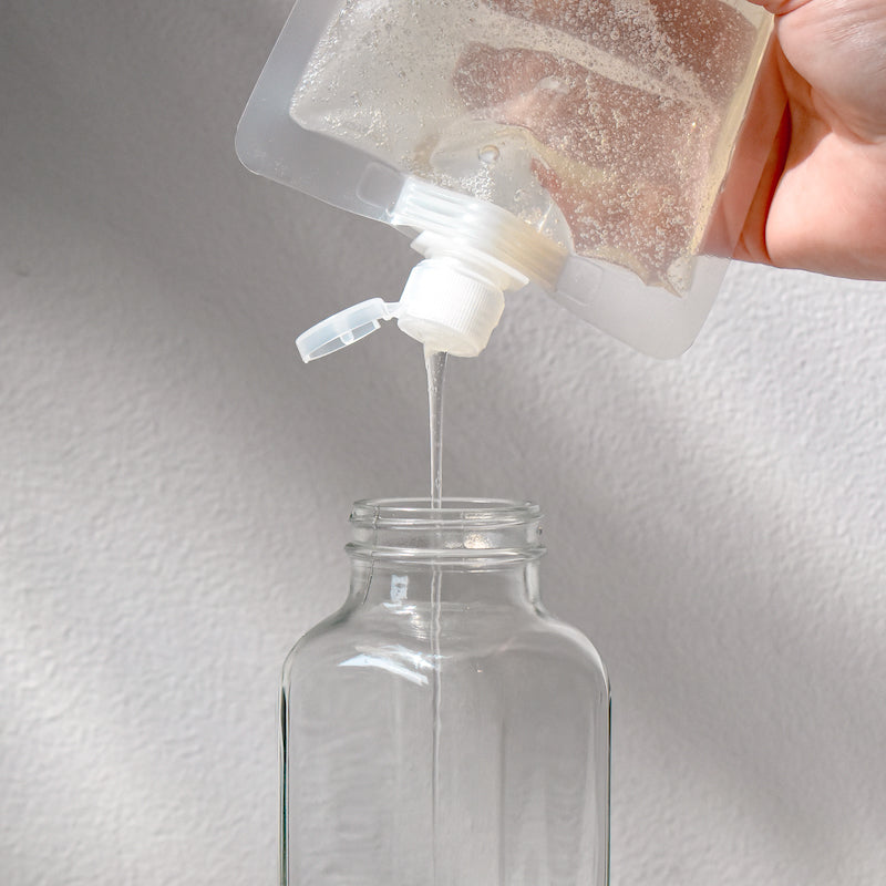 A hand pours Rustic Strength Body Wash from a plastic refill pouch into a clear glass bottle, showcasing natural hydration against a simple light backdrop.