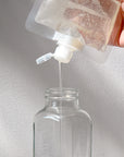 A hand pours Rustic Strength Body Wash from a plastic refill pouch into a clear glass bottle, showcasing natural hydration against a simple light backdrop.