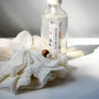 A clear bottle labeled Bubble Bath rests on a white surface beside the Bamboo Switch Shower Pouf, an eco-friendly, plant-based bath accessory with a wooden bead detail, all softly lit by natural sunlight.