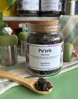 A refillable jar of Pu'erh tea. The jar has a cork lid and an attachable bamboo spoon. the spoon is in front full of tea.