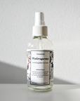 A clear Carina Organics Hairspray bottle with a white label and cap sits on a white surface, casting a shadow. The label displays usage instructions and delicate floral illustrations along the sides.