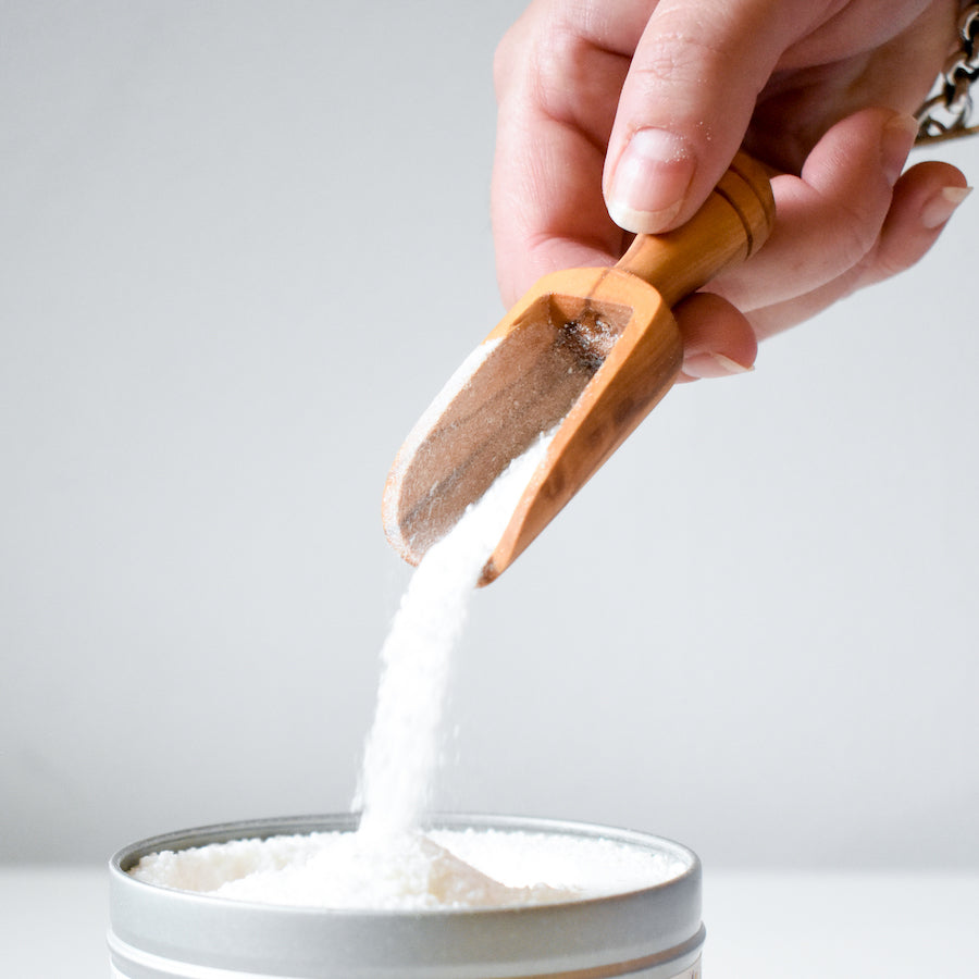 A hand pours white powder with the Vesper & Vine Wood Scoop into a gray metal container against a plain white background.