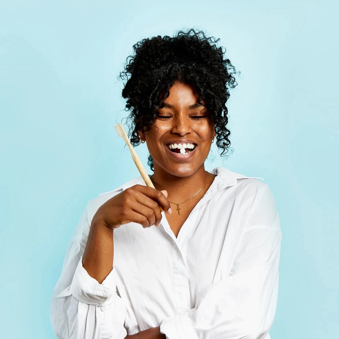 A smiling woman with curly hair in a white shirt holds Huppy Foaming Tooth Tabs, showing the product on her teeth against a light blue background.