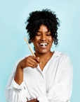 A smiling woman with curly hair in a white shirt holds Huppy Foaming Tooth Tabs, showing the product on her teeth against a light blue background.