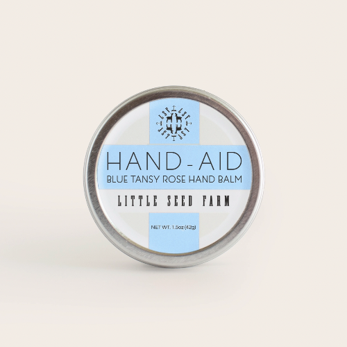 A round metal tin labeled Healing Hand Balm by Little Seed Farm is shown against a plain background. This moisturizing balm, made with organic ingredients, features a white label with blue accents and black text.