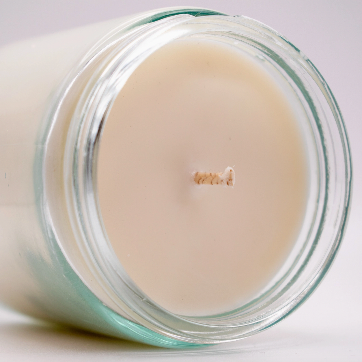 Close-up of an unlit Wood Candle Co. Soy Candle, hand-poured white soy wax in a clear glass jar lying on its side with a centered wick visible. The softly lit background is plain.