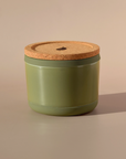 The Arbor Made Refillable Candle Jar by Arbor Made is a green cylindrical container with a cork lid and silicone bottom, ideal for use with wax refills, shown on a beige background casting a soft shadow to the right.