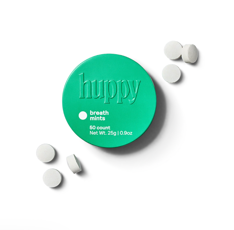 A green circular tin of Huppy Breath Mints sits on a white background, with several round white mints scattered nearby. The sugar-free, naturally flavored tin contains 50 mints and weighs 25g (0.9oz).