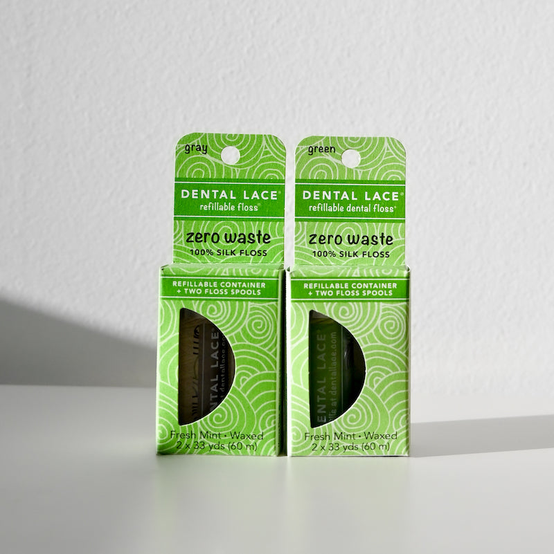 Two boxes of Dental Lace Compostable Floss in eco-friendly green packaging, labeled “zero waste” and “100% silk,” sit side by side on a white surface, featuring compostable silk floss in bright natural light.