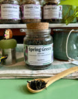 A glass jar of Spring Green tea with a cork lid sits on a green cloth, with a wooden spoon holding loose tea leaves in front. Other tea jars and a ceramic mug are blurred in the background.