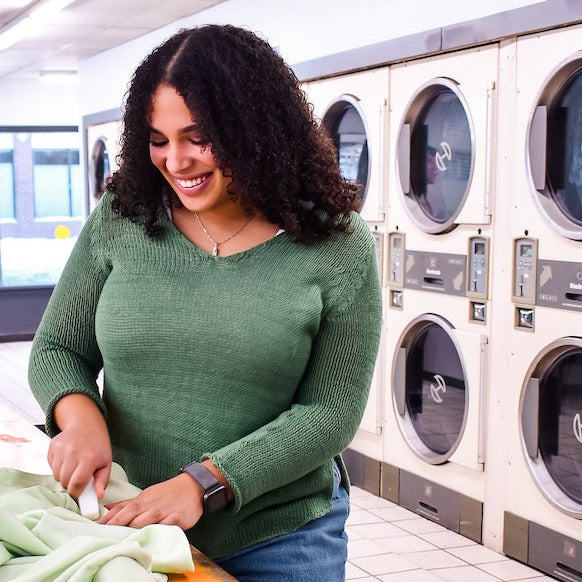 A woman with curly hair in a green sweater and jeans smiles as she uses the Meliora Laundry Stain Stick to treat toddler clothes at a laundromat, with large dryers in the background.