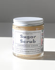 A glass jar of Shade Tree Naturals Sugar Scrub with a silver lid sits on a white surface. The label features floral designs and usage instructions, highlighting its gentle exfoliation with organic sugar.