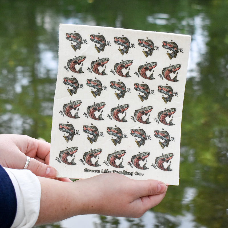 A person holds a washable Swedish Dishcloth by Potluck Press with colorful fish illustrations, trees and water blurred behind it. The text Green Line Trading Co. appears at the bottom of this super absorbent dishcloth.