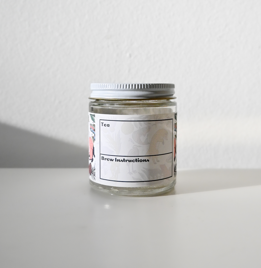 A small glass jar with a white lid from Loose Leaf Tea Co. sits on a white surface. The label reads Peppermint Herbal Tea with brew instructions, suggesting a classic and refreshing tea experience, set against a plain, light-colored wall.