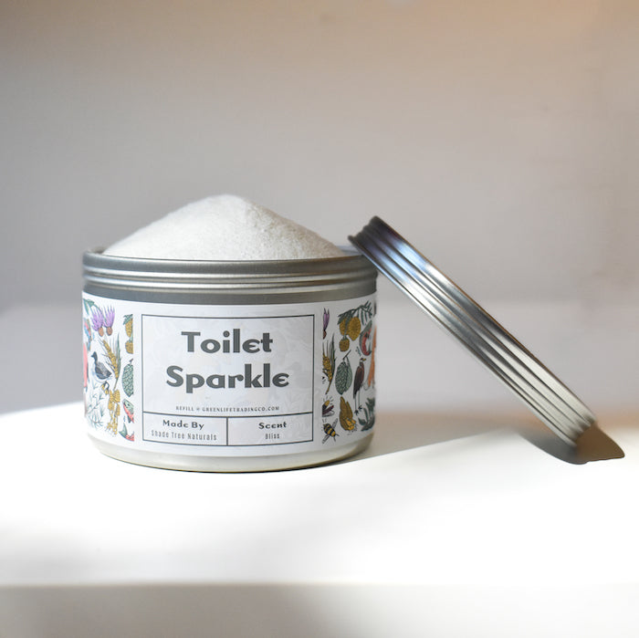 A round, silver tin of Shade Tree Naturals Toilet Sparkle sits in soft natural light, lid partly open to reveal white powder inside; the label features floral designs and the product name.