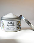 A round, silver tin of Shade Tree Naturals Toilet Sparkle sits in soft natural light, lid partly open to reveal white powder inside; the label features floral designs and the product name.