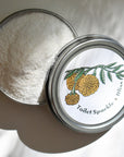 A round metal tin of white powder is displayed with a partially open lid showing yellow flowers, green leaves, and the text Toilet Sparkle by Shade Tree Naturals.