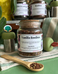 A refillable jar of Vanilla Rooibos tea. The jar has a cork lid and an attachable bamboo spoon. The spoon is in front of the jar with vanilla rooibos tea in it.