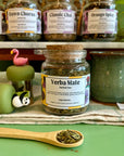A refillable glass jar of Yerba Mate herbal tea with a cork lid sits on a striped cloth on a green surface, with a wooden spoon of loose tea in front and other tea jars in the background.