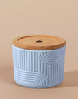 The Arbor Made Refillable Candle Jar in the silicone jar style and in the color sky. The jar features a cork lid with a small leaf design on it and a silicone body with a linear geometric pattern on it. The Jar is siting on a beige background.