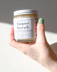 A hand with green-painted nails holds a Shade Tree Naturals Sugar Scrub jar under bright light, displaying some of the gently exfoliating sugar scrub on their hand.