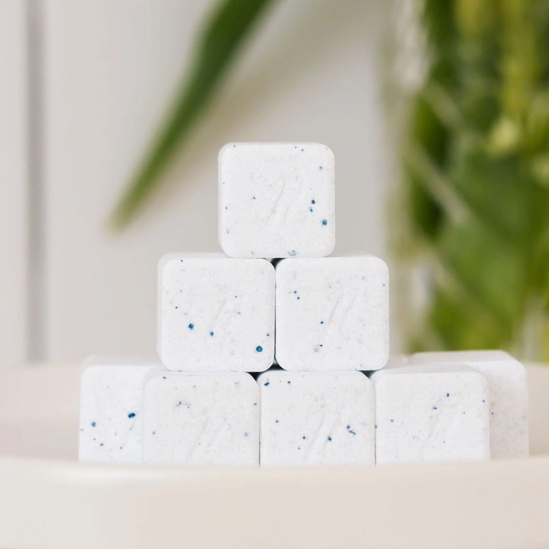 A small stack of Nellies Dishwasher Cube tablets—white squares with blue specks—sits on a white surface, with a blurred green plant in the background.