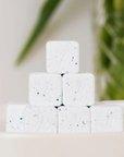 A small stack of Nellies Dishwasher Cube tablets—white squares with blue specks—sits on a white surface, with a blurred green plant in the background.
