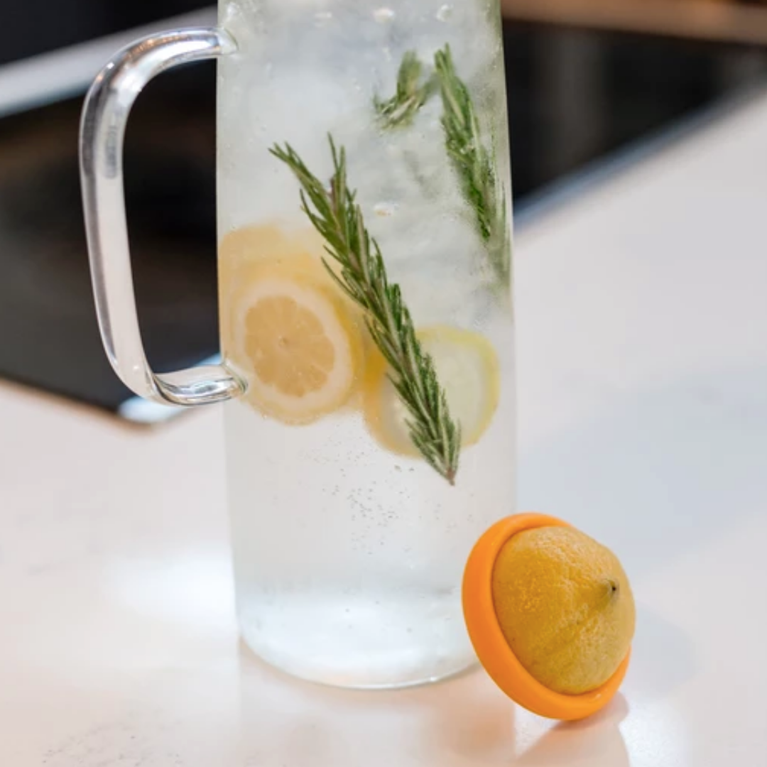 A glass pitcher of water with lemon slices and rosemary sits on a countertop. In front, a lemon half in a yellow citrus squeezer is kept fresh with Food Huggers by Food Huggers—an ideal way to reduce food waste.