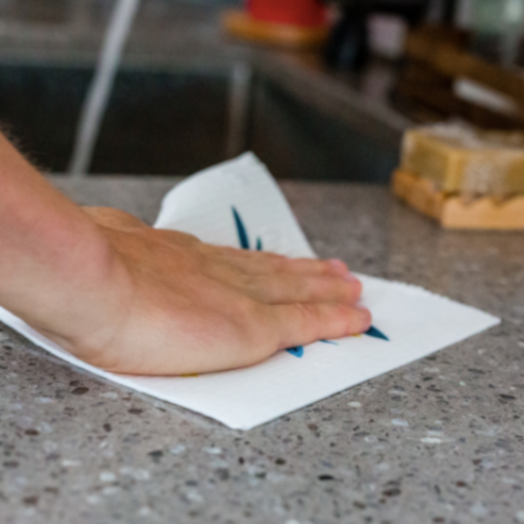 A hand wipes a gray speckled kitchen countertop with a Potluck Press Swedish Dishcloth. Sponges and a sink are visible in the background.