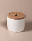 The Arbor Made Refillable Candle Jar by Arbor Made is a small white ceramic container with a round cork lid featuring a Leaf symbol and a silicone bottom—ideal for use as refillable candle jars with your favorite wax refills.