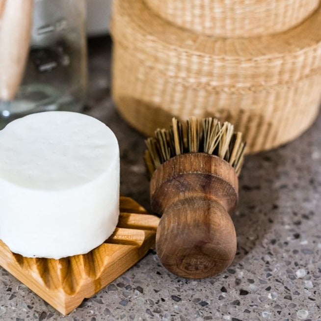 A round white soap rests on a wooden dish next to the Zefiro Pot Brush, featuring natural bristles and a beechwood handle, displayed on a speckled countertop with woven baskets in the background.
