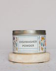 A metal tin labeled Nellies Dishwasher Powder, made with a biodegradable plant-based formula, sits on a round stone slab. The white label features botanical illustrations and greenlifetradingco.com below the main text.