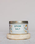 A tin of Salt Works Epsom Salt, labeled pharmaceutical-grade and decorated with botanical illustrations, sits on a round stone coaster against a light background, evoking classic bath products.