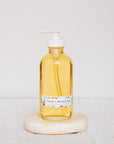 A clear pump bottle of yellow Green Life Trading Co. Hair, Beard & Body Oil sits on a round stone coaster against a light background.