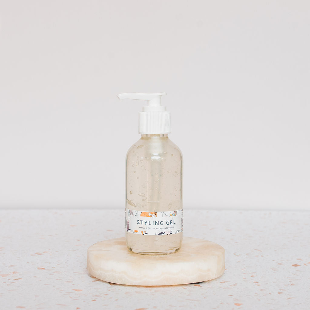 A clear Glass Pump Bottle by Container sits on a round, light-colored stone against a pale background. The labeled bottle contains clear gel and features a white pump top—ideal for your next DIY creation.