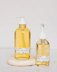 Two clear glass pump bottles of Green Life Trading Co. Hair, Beard & Body Oil, enriched with almond oil, rest on a round stone platform against a light, neutral background.