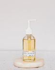 A clear glass pump bottle labeled Hair, Beard & Body Oil by Green Life Trading Co., enriched with jojoba oil and filled with yellow oil, sits on a round stone slab against a light, neutral background.