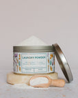 A tin of Meliora Laundry Powder with a floral label sits on a stone slab. Its lid leans to the side, and a wooden scoop filled with MADE SAFE Certified powder rests in front of the container.