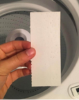 A hand holds a white plantbased Laundry Detergent Strip in front of an open washing machine drum.