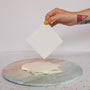 A hand with yellow nail polish and geometric tattoos holds a square sheet of TruEarth Laundry Detergent Strips above a stack of similar sheets on a round, marble-patterned surface.