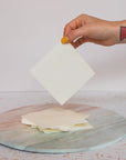 A hand with yellow nail polish and geometric tattoos holds a square sheet of Plant based Laundry Detergent Strips above a stack of similar sheets on a round, marble-patterned surface.
