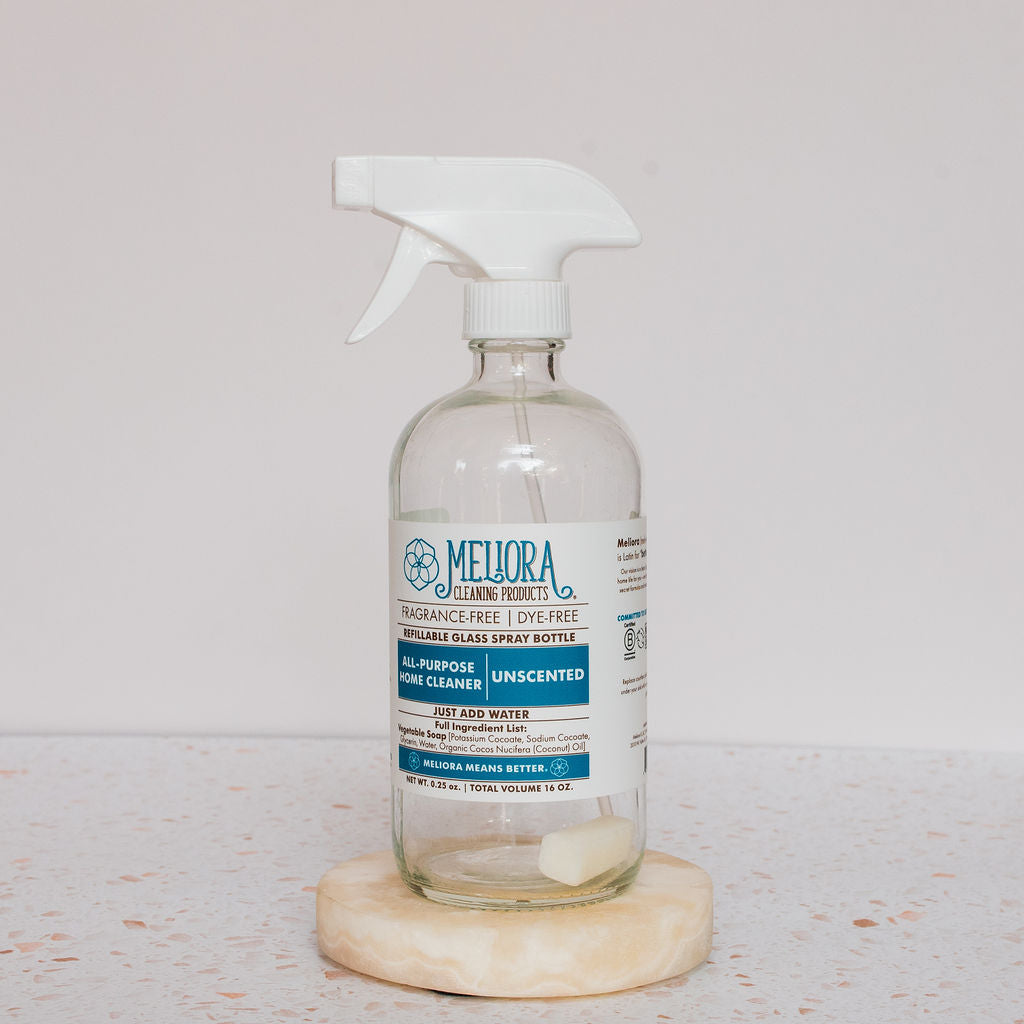 A clear glass spray bottle with a white trigger and blue-and-white label reads Meliora All-Purpose Home Cleaner Tabs, Unscented. It sits on a round stone coaster alongside an unscented tab for hard surface cleaning.