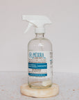 A clear glass spray bottle with a white trigger and blue-and-white label reads Meliora All-Purpose Home Cleaner Tabs, Unscented. It sits on a round stone coaster alongside an unscented tab for hard surface cleaning.