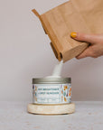 A hand with yellow-painted nails pours white powder from a brown paper bag into a labeled container of Meliora Oxy Brightener & Spot Remover, which rests on a circular stone atop a speckled surface.