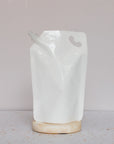 A plain white stand-up pouch of Taylors Naturals Baby Shampoo & Body Wash with a spout rests upright on a round beige stone against a light, neutral background.