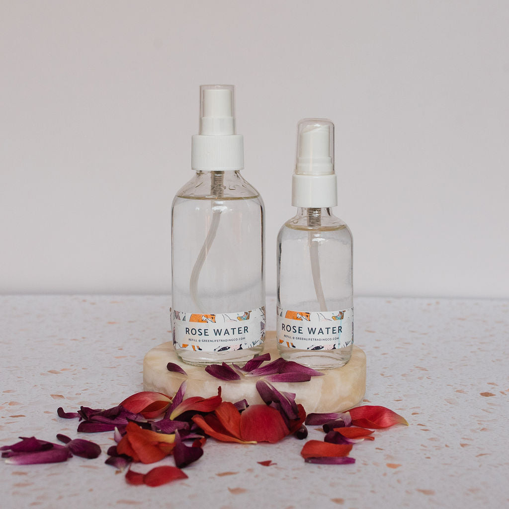 Two Glass Misting Bottles from Container sit on a round stone coaster amidst red and pink rose petals on a light, speckled surface. Each bottle releases a refreshing fine mist, displayed against a simple background.