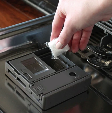 A hand places a Dropps Lemon Dish Pod with a biodegradable formula into the detergent compartment of an open dishwasher door.