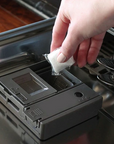A hand places a Dropps Lemon Dish Pod with a biodegradable formula into the detergent compartment of an open dishwasher door.