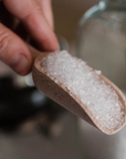 A hand holds a small wooden scoop filled with coarse, white Salt Works Epsom Salt, with a glass container blurred in the background.